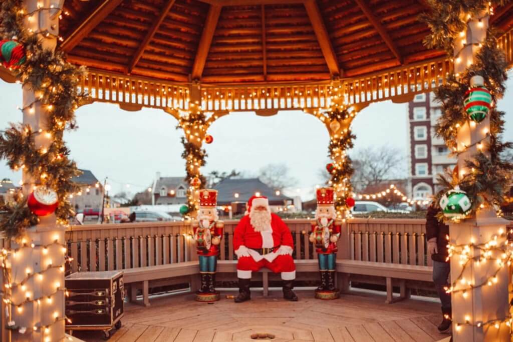 Montauk gazebo lit up with warm white christmas lights, garland, green and red ornaments, and toy soldiers leading the way to a jolly Santa Claus sitting inside