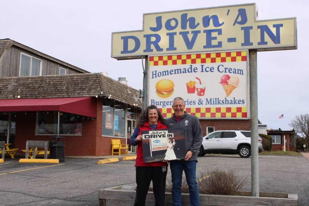 Maureen and David Rutkowski holding their wedding photo in front of the John's Drive In sign then, in front of John's Drive-In today
