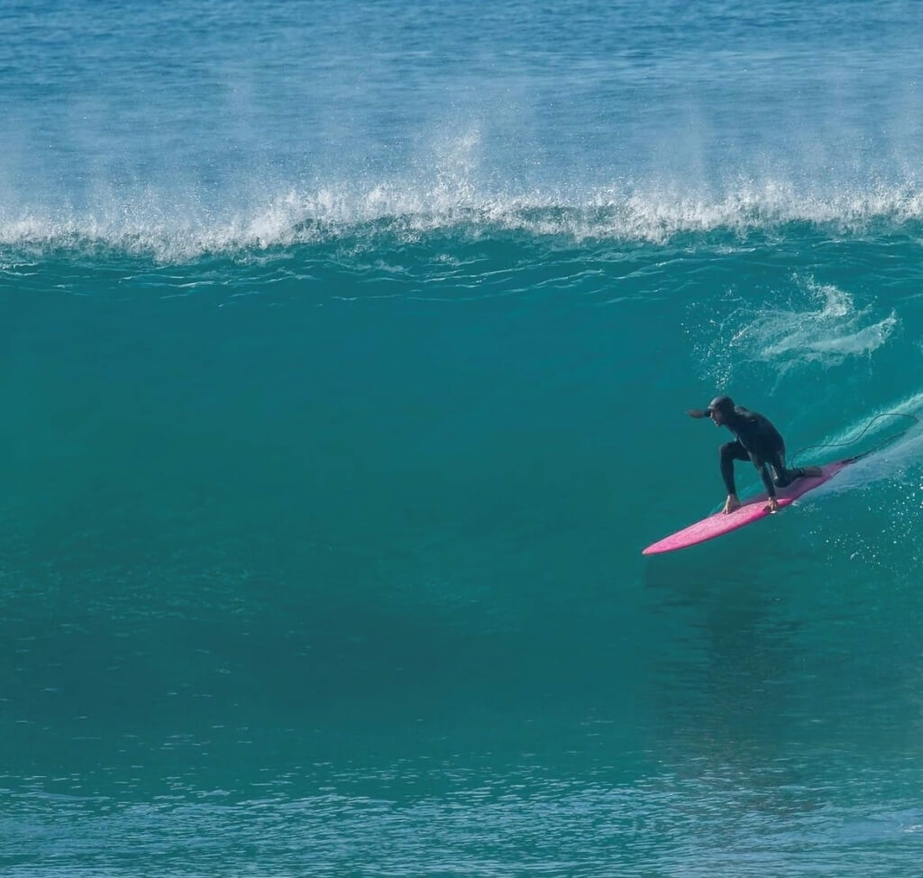 Surfer in head-to-toe wetsuit on pink surfboard rides a turquoise wave off of Montauk, courtesy of Coreyswave