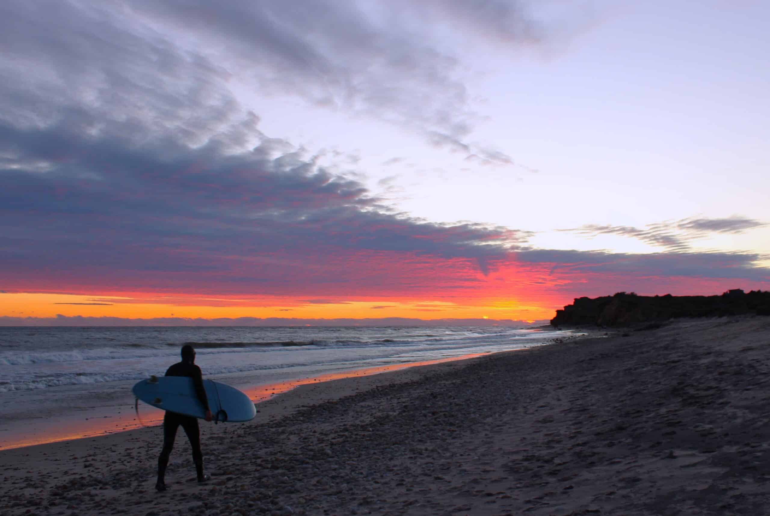Surfer in full-body wetsuit and cap carrying surfboard into a purple, pink, and orange sunset over Ditch Plains Beach, November 2024