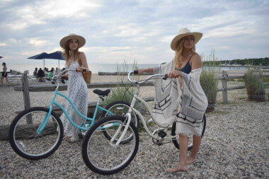 Two bohemian style blonde women with cruiser bicycles pulling up to Navy Beach Restaurant; stunning sea views of Fort Pond Bay expand behind them