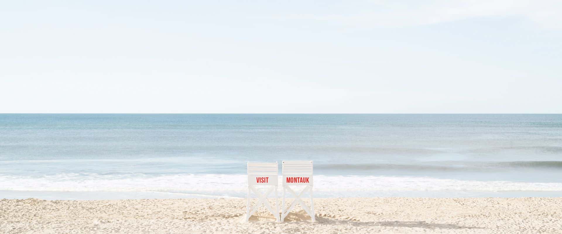 "Visit Montauk" painted on the back of two lifeguard chairs on a beach.