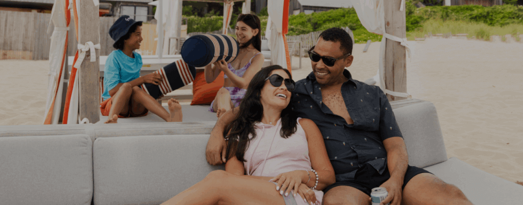 Smiling couple lounging on the beach at Gurney's Montauk Resort with two kids playing in the background