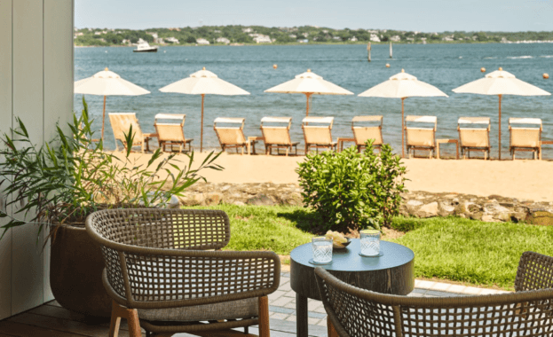 'Image of the luxurious Montauk Yacht Club water front, featuring a row of lounge chairs and umbrellas on the beach and shaded patio furniture.